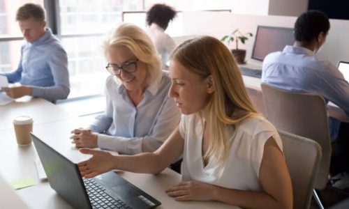 Young saleswoman manager mentor advisor consulting old female client or new employee pointing at laptop showing business presentation teaching instructing at meeting in modern corporate shared office